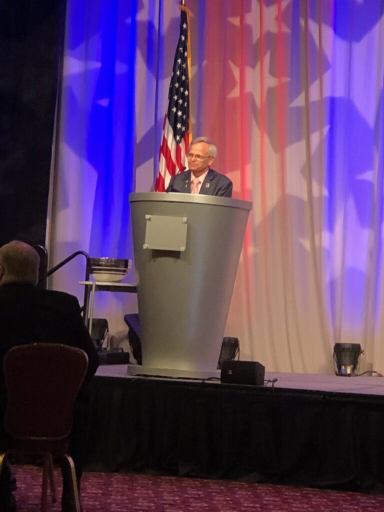 A speaker stands at a podium decorated with an American flag backdrop