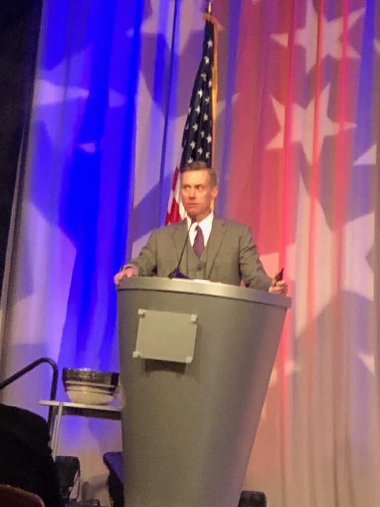 A speaker in a gray suit addresses an audience from a modern podium