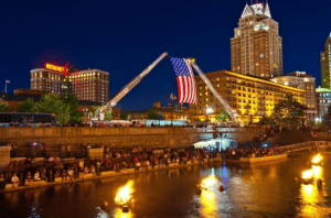 A large American flag is displayed between firetruck ladders above a waterfront